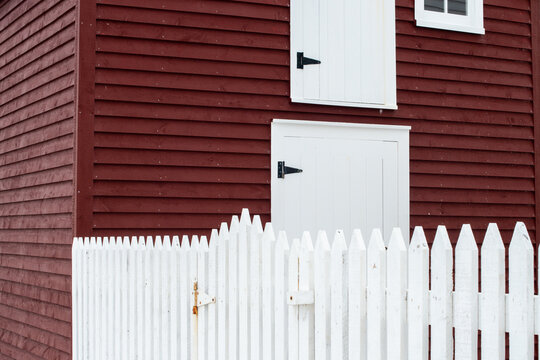 The exterior of a red wooden cape cod clapboard or batten board siding wall of a country style barn with a white wooden picket fence in the foreground. There are two doors and a window in the building