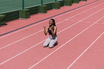 Happy African American woman in sportswear sits on terracotta track of stadium rejoicing with success in long-distance race. Young professional female athlete enjoys taking part in running competition