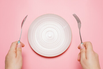 Female hands holding a fork and a spoon next to an empty plate on a pink background close-up top...