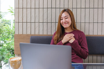 A beautiful young asian woman holding hands on chest while using laptop computer for video call,...