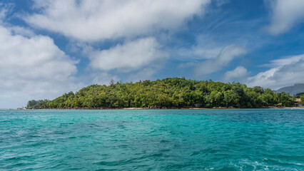 A tropical island in the ocean is completely covered with lush green vegetation. Aquamarine water and blue sky with clouds. Seychelles