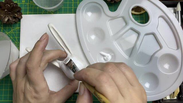 Master paints clay rabbit with white acrylic. Plastic palette, bunny figure in female hands on table. Leisure and craft in quarantine.