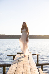 Beautiful Russian girl dressed in a white dress, walking along a wooden pier on the bank of a river or a lake