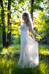 Beautiful young Russian girl dressed in a white dress, walks through the forest among the oaks in the rays of the sunset