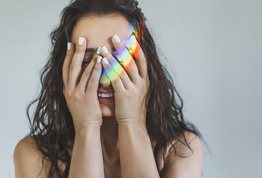 Portrait of a young woman covering her eyes with her palms and laughing with the reflection of a rainbow on her hands. The girl closed her eyes and enjoys life. Emotional psychological state. lgbt sym