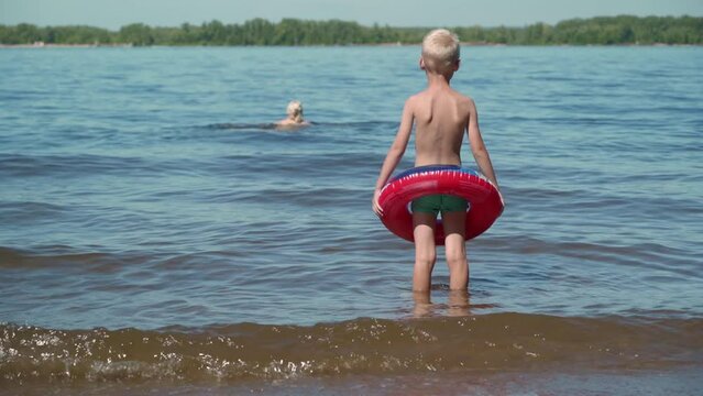 A hot summer day is a great option for a family holiday on the lake. A little boy watches as his mother swims away from him into the middle of the pond. The child learns to swim with a life buoy.