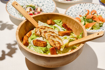 Classic caesar salad with grilled chicken and crouton in wooden bowl on white table. Caesar salad with sauce and wooden spoon. Italian lunch with caesar salad on sunny day. Summer menu.
