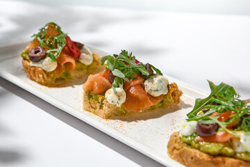 Aesthetic composition with salmon bruschetta on white background with shadows from flowers. Italian bruschetta with salmon, avocado, cheese and olives on fine dining in summer. Elegant menu concept.