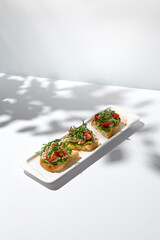 Aesthetic composition with shrimp bruschetta on white background with shadows from flowers. Italian bruschetta with shrimp, tomatoes and arugula on fine dining in summer. Elegant menu concept.