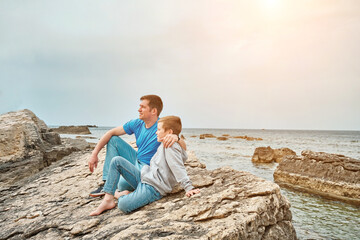 Fototapeta na wymiar Happy family, father and son bonding, sitting on stone by the sea looking at view enjoying summer vacation. Togetherness Friendly concept 