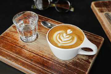One cup of cappuccino with latte art. Coffee in ceramic cup with glass of water on brown wooden table in cafe.