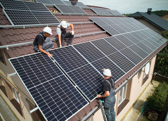 Men installers mounting photovoltaic solar moduls on roof of house. Engineers in helmets installing...