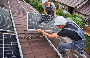 Men technicians carrying photovoltaic solar moduls on roof of house. Engineers in helmets building...