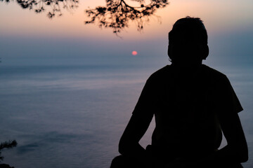 silhouette of a young man sitting next to a pine tree with the sunset and the sea in the background