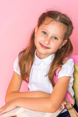 happy smiling little girl in uniform with a backpack and books poses on pink background in studio. schoolgirl rejoices at beginning of classes, goes to study with pleasure. Back to school. Close up