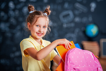 Cute happy schoolgirl puts stationery in a backpack. Preparation for school. Back to school. A little girl is learning to self-assemble a school backpack.