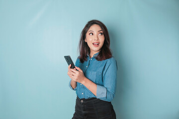 Naklejka premium A portrait of a happy Asian woman is smiling and holding her smartphone wearing a blue shirt isolated by a blue background