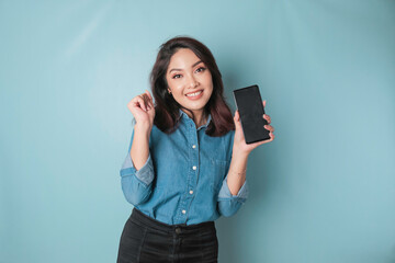 Naklejka premium A young Asian woman with a happy successful expression wearing a blue shirt holding smartphone isolated by blue background