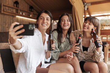 Happy young caucasian women take selfie on phone enjoying drink on weekend. Brunettes wear casual...