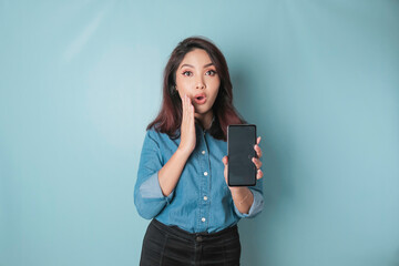 Surprised Asian woman wearing blue shirt pointing at her smartphone, isolated by blue background © Reezky
