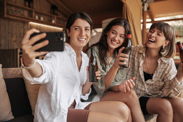 Three cheerful young caucasian ladies with beer take photo on smartphone spending leisure time at party. Brunettes smile, wear casual clothes. Happy weekend concept.