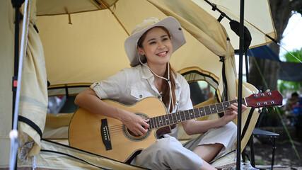 Fototapeta na wymiar Happy young Asian woman playing guitar in camping tent. Outdoor holidays summertime concepts