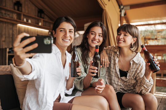 Positive young caucasian girlfriends are photographed on smartphone, relax with beer in campsite. Brunettes smile, wear shirts and shorts. Lifestyle concept