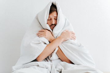 Adult frozen man wrapped in a blanket and hugging himself while sitting on a bed indoors. Lonely guy with closed eyes warming in a cold house - obrazy, fototapety, plakaty
