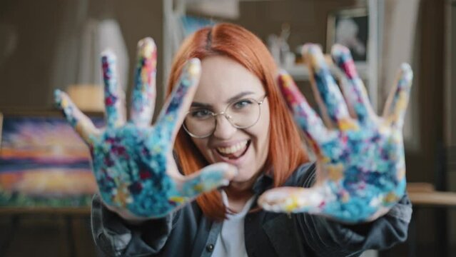 Happy funny Caucasian red-haired woman in eyeglasses girl painter wear glasses looking at camera posing in art studio laughing show palms dirty soiled in acrylic paints hands after painting drawing