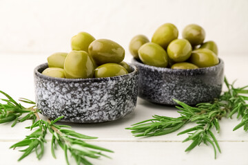 Bowls of tasty green olives and rosemary on light wooden table, closeup