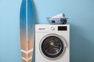 Basket with towels on washing machine and surfboard near blue wall