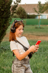 A teenage girl stands in a park with a smartphone in her hands. A 12-year-old girl listens to music on wireless headphones.
