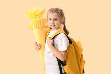 Cute little girl with yellow school cone and backpack on beige background
