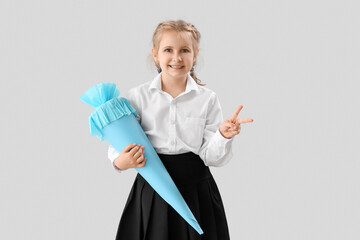 Cute little girl with blue school cone showing victory gesture on light background