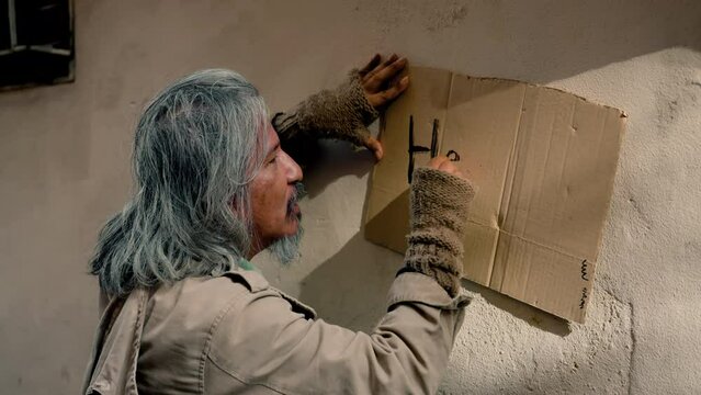 4K, Close Up A homeless old man with long hair dressed in dirty clothes is writing a note on a piece of paper asking for help from a passerby, he has no home and no job and has not eaten in days.