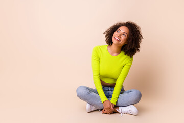 Full size photo of nice young woman curly hair folded legs look copyspace dressed stylish yellow outfit isolated on beige color background