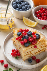 Stack of French crepes or Russian blini on white plate on light background. Thin pancakes with raspberries, blueberries, currant, icing sugar, lemon jam and tea. Selective focus.