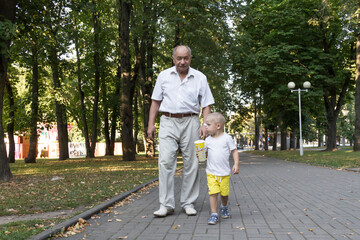 Grandfather and grandson are walking in the park along the path with a glass of popcorn. An old man and a little boy have snacks on the go.