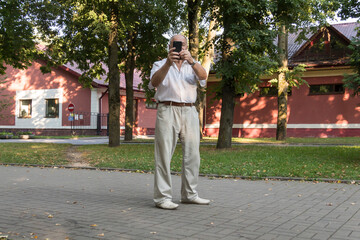 An elderly man walks alone in the park in the summer. A modern pensioner, businessman in a white shirt and trousers takes pictures with a camera in a mobile phone