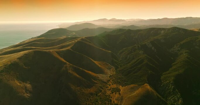 Tremendous rocky hills in the rays of bright sun. Mountainous landscape nearby the ocean at the backdrop of orange sky. Aerial perspective.