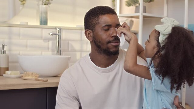 Tilt up slowmo of little girl sitting on father laps in bathroom while applying eye patches on his face