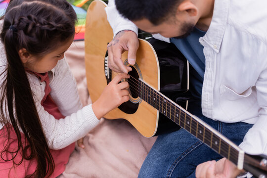 Preteen asian child playing acoustic guitar with father on blanket in park.