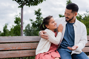 Smiling asian dad hugging and talking to daughter on bench in park.