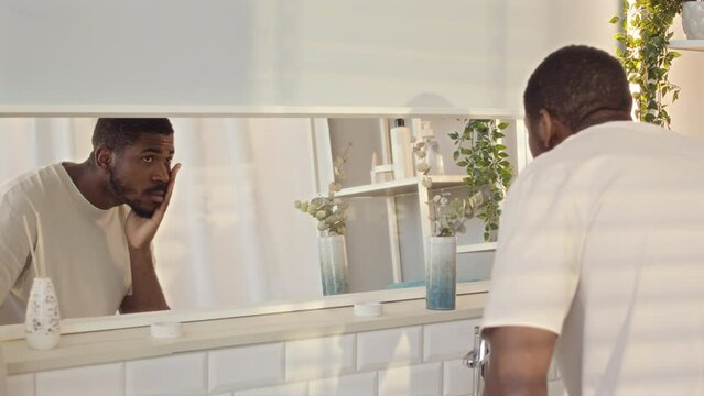 Medium slowmo of brutal young Black man examining his reflection in mirror at modern minimalist style bathroom