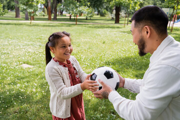 Smiling asian man holding soccer ball near daughter in summer park.