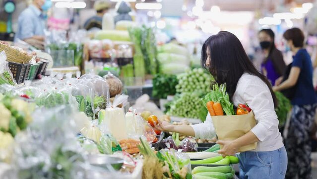 Asian woman buying vegetables and puts tomato in paper shopping bag. Lots of Vegetables in shelves at fresh market and Blurred people are shopping in background. Recycled paper shopping bags.