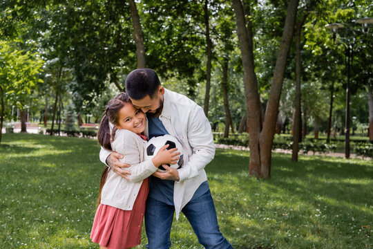 Asian father hugging daughter with football in park.