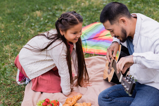 Cheerful asian girl looking at father playing acoustic guitar near fruits and croissants on blanket in park.