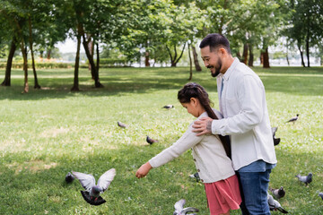Asian dad hugging daughter near birds in summer park.
