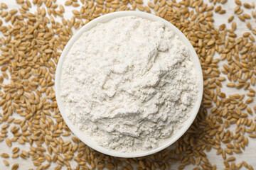 Bowl with organic flour and grains of wheat on white table, top view